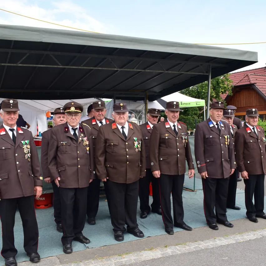 Eine Gruppe von Männern in braunen Uniformen steht unter einem Zelt, trägt Hüte und Medaillen. Dahinter stehen Zelte, Bäume und ein Haus mit rotem Dach. Die Männer stehen in einer Reihe.