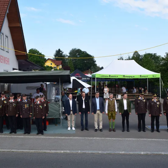 Eine Gruppe uniformierter Männer und Beamter steht in einer Reihe draußen, mit Zelten und einem Gebäude im Hintergrund.