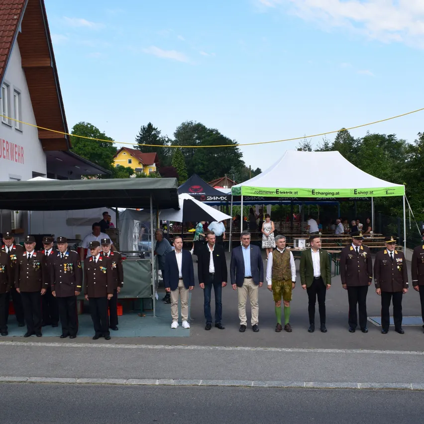 Eine Gruppe uniformierter Männer und Beamter steht in einer Reihe draußen, mit Zelten und einem Gebäude im Hintergrund.