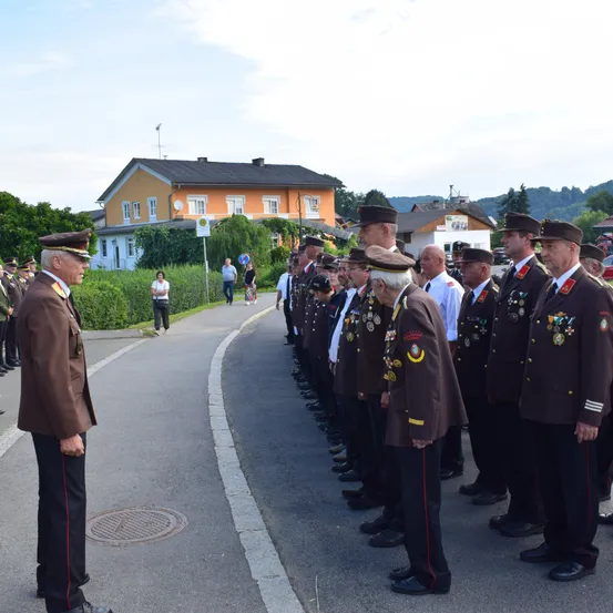 Eine Reihe von Militärangehörigen in Uniform steht am Straßenrand, gegenüber einem Mann in Uniform. Gebäude und Grünflächen sind im Hintergrund zu sehen.