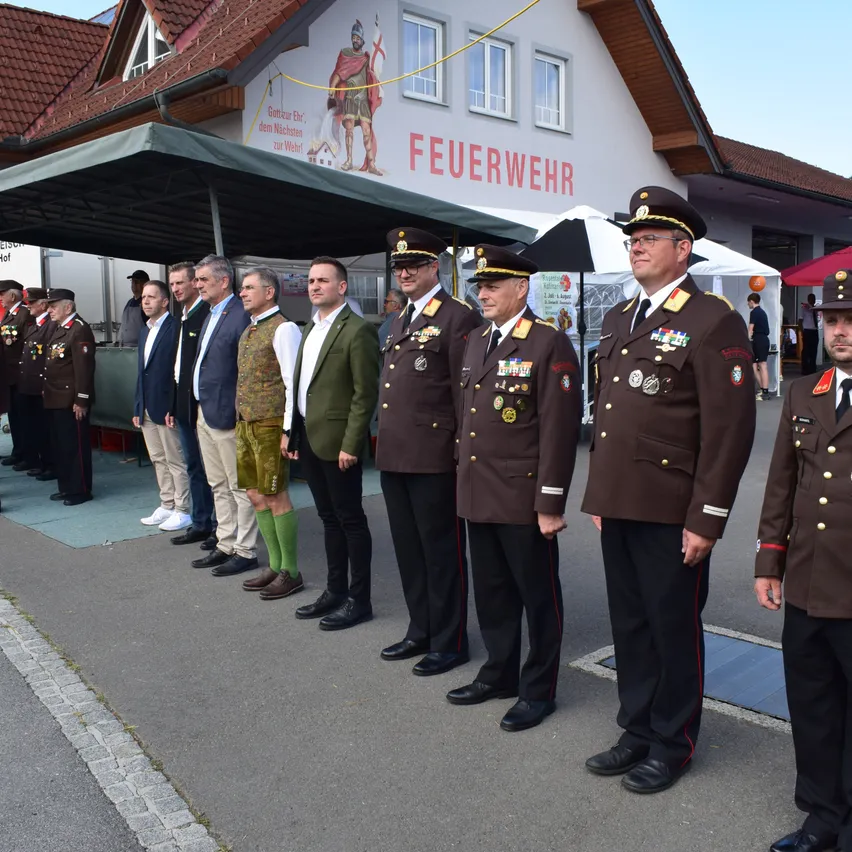 Eine Reihe von Militärangehörigen und Beamten steht vor einem Gebäude mit dem Schild 'Feuerwehr', einem Vordach und Zelten dahinter.