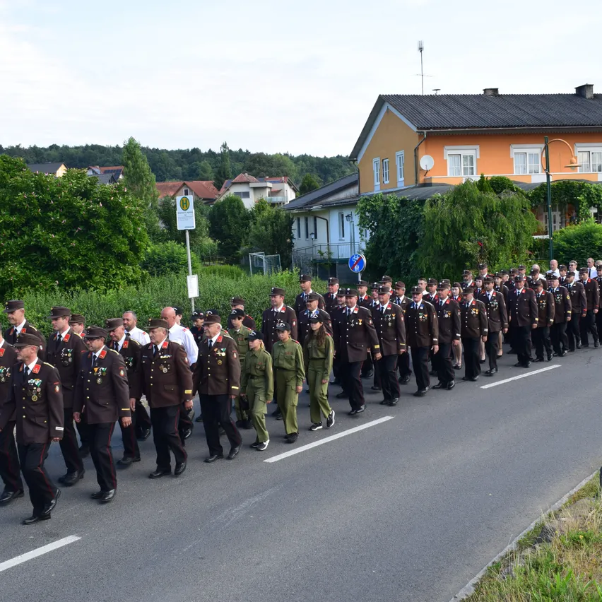 Ein Militäraufmarsch mit Soldaten in Uniform, die eine Straße hinuntermarschieren, mit Häusern und Grünflächen im Hintergrund.
