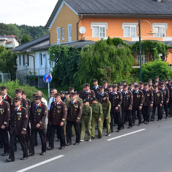 Eine große Gruppe uniformierter Personen, wahrscheinlich Militärangehörige, marschiert in einer Reihe auf einer Straße. Sie tragen Mützen und Medaillen. Hinter ihnen sind Gebäude mit Balkonen und ein Fahrradschild zu sehen.