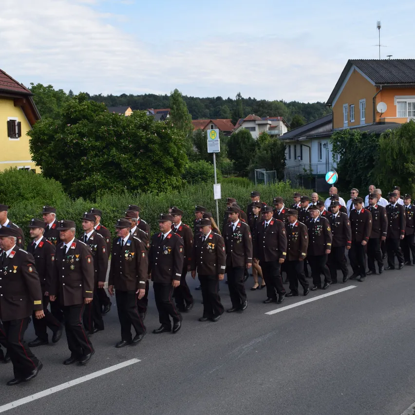 Eine Gruppe uniformierter Personen marschiert eine Straße hinunter, mit Wohngebäuden und Grünflächen im Hintergrund.