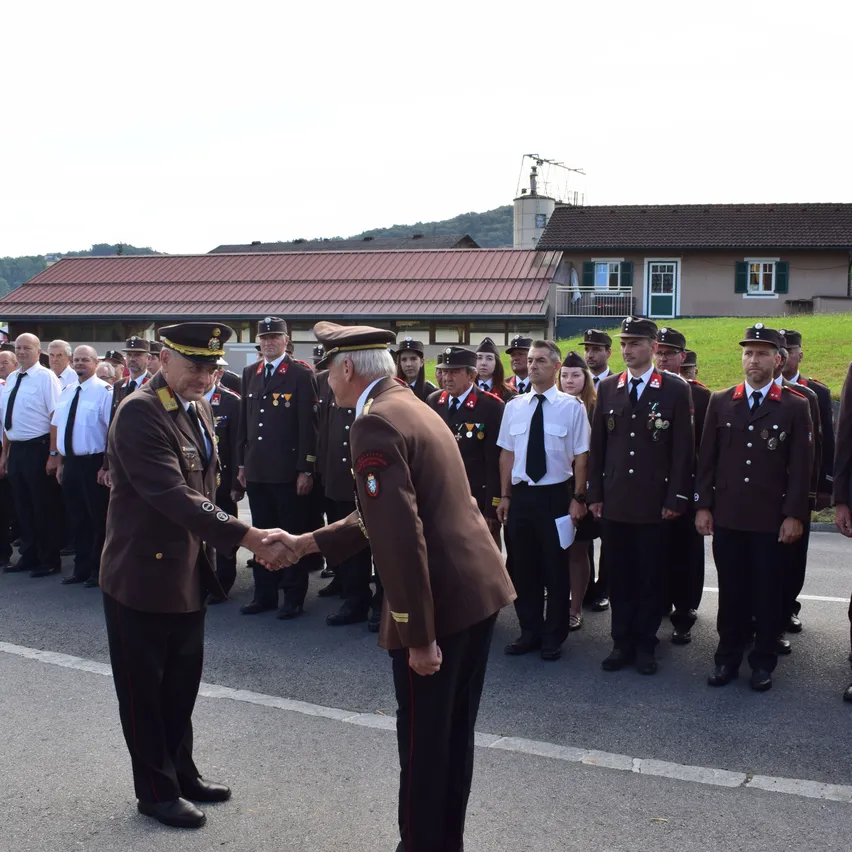 Zwei Männer in Militäruniformen schütteln sich die Hände vor einer Menschenmenge. Sie stehen auf einem Außengelände, möglicherweise einer Militärbasis. Die Menge steht in Reihen, und einige tragen Hüte. Die Männer tragen Medaillen und Dekorationen an ihren Uniformen.