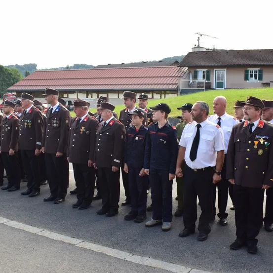 Eine Gruppe uniformierter Personen, darunter Militärangehörige und Zivilisten, steht in einer Reihe auf einer Straße, mit einem Haus und Grünflächen im Hintergrund.