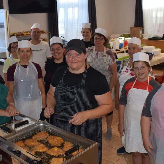 Eine Gruppe von Menschen in weißen Schürzen und Mützen steht in einer Küche und kocht Essen in einer Fritteuse.