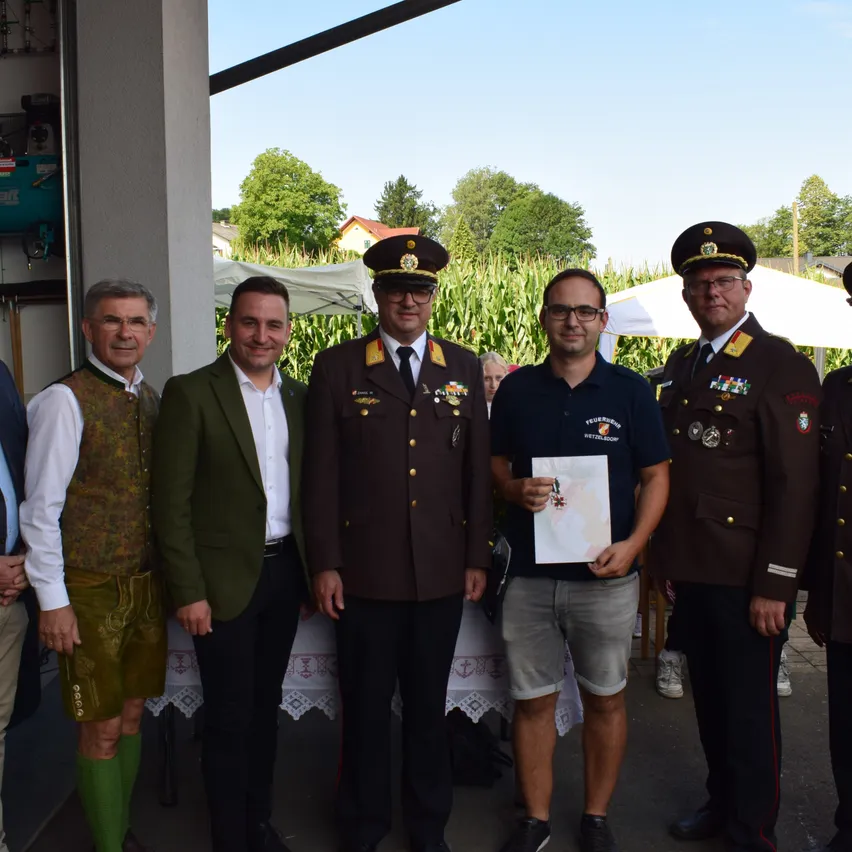 Fünf Männer stehen unter einem Vordach, zwei in Militäruniformen und drei in ziviler Kleidung. Die zentrale Figur hält eine Urkunde, mit einem lächelnden Mann auf seiner linken Seite und einem Mann in einer braunen Uniform auf seiner rechten Seite. Bäume und ein Haus sind im Hintergrund.