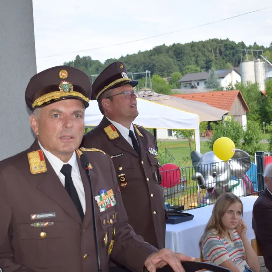 Zwei Männer in Militäruniformen sprechen bei einer Veranstaltung im Freien. Hinter ihnen steht ein Zelt und ein Mädchen sitzt an einem Tisch. Ein gelber Luftballon und eine Elefantenstatue sind im Hintergrund.