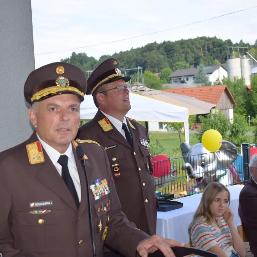 Zwei Männer in Militäruniformen sprechen bei einer Veranstaltung im Freien. Hinter ihnen steht ein Zelt und ein Mädchen sitzt an einem Tisch. Ein gelber Luftballon und eine Elefantenstatue sind im Hintergrund.