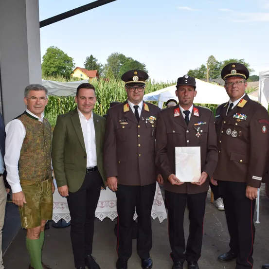 Fünf Männer stehen vor einem Gebäude. Drei von ihnen tragen Militäruniformen und einer hält eine Auszeichnung. Der Mann auf der linken Seite trägt ein traditionelles Outfit. Der Mann in der Mitte trägt einen grünen Anzug.