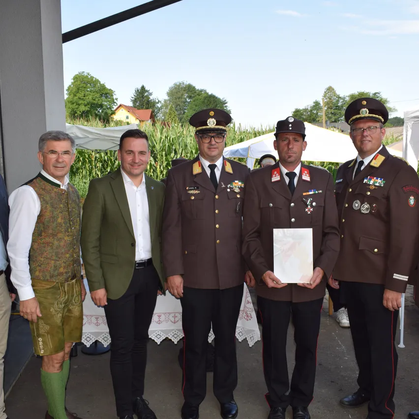 Fünf Männer stehen vor einem Gebäude. Drei von ihnen tragen Militäruniformen und einer hält eine Auszeichnung. Der Mann auf der linken Seite trägt ein traditionelles Outfit. Der Mann in der Mitte trägt einen grünen Anzug.