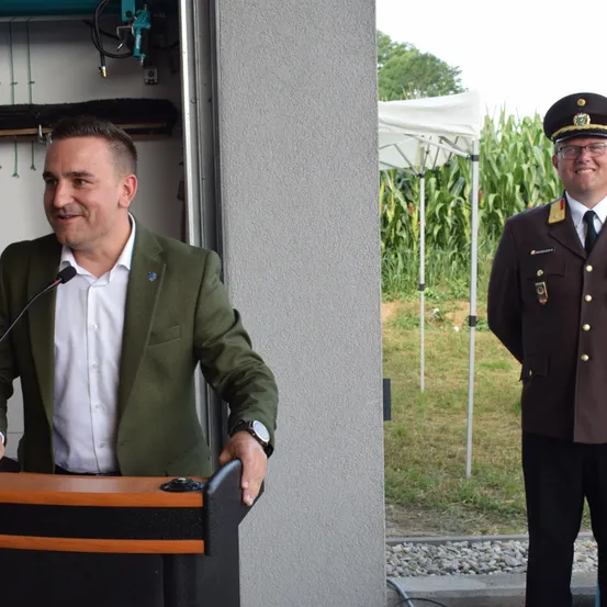 Ein Mann steht mit einem Mikrofon am Rednerpult, trägt einen grünen Anzug. Hinter ihm steht ein uniformierter Mann neben einem Zelt in einem Grasfeld.