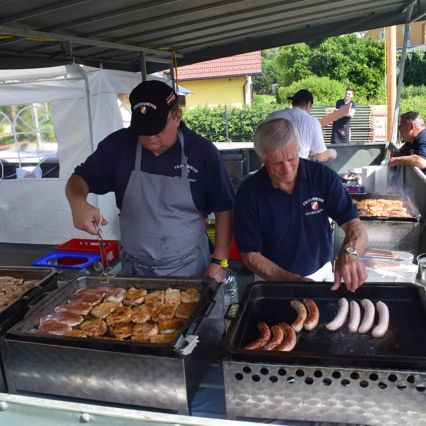 Zwei Männer grillen Würstchen und Pfannkuchen auf Grills bei einer Outdoor-Veranstaltung. Ein Zelt und mehrere Personen sind im Hintergrund.