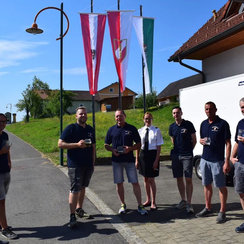 Eine Gruppe von Menschen in Uniformen, die Gläser halten, steht draußen in der Nähe eines weißen Lieferwagens mit Flaggen und einem Gebäude. Das Gebiet hat eine Straße, einen Bürgersteig, Straßenlaternen und einen grasbewachsenen Hügel.