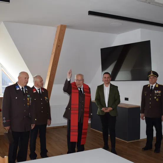 Fünf Männer in Uniformen stehen in einem Raum. Ein Mann in der Mitte trägt einen roten Schal. Hinter ihnen ist ein Fernseher an der Wand montiert. Links ist ein Holzbalken zu sehen.