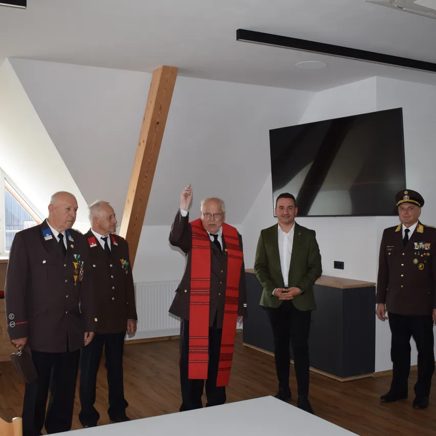 Fünf Männer in Uniformen stehen in einem Raum. Ein Mann in der Mitte trägt einen roten Schal. Hinter ihnen ist ein Fernseher an der Wand montiert. Links ist ein Holzbalken zu sehen.