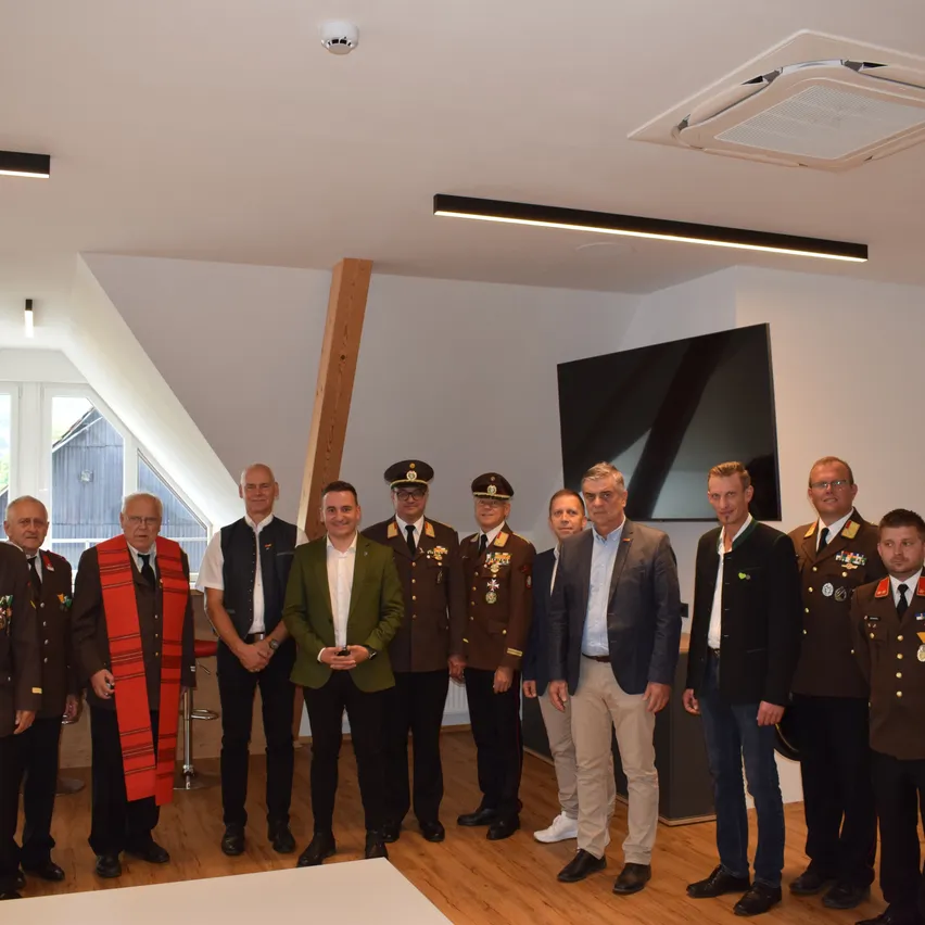 Mehrere Männer in Militäruniformen stehen in einem Raum mit Holzboden, lächeln und posieren für ein Foto. Ein Mann in einem grünen Mantel sticht hervor. Hinter ihnen ist ein großer Fernseher an der Wand montiert.