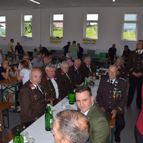 Eine Gruppe von Männern in Uniformen sitzt an einem Tisch in einer Halle. Einige stehen in der Nähe der Tische. Auf dem Tisch befinden sich Flaschen, Gläser und Blumen. Die Halle hat weiße Wände und Fenster.