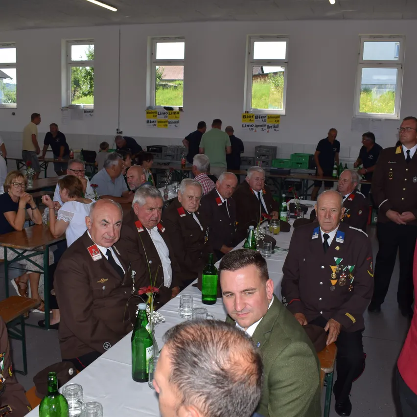 Eine Gruppe von Männern in Uniformen sitzt an einem Tisch in einer Halle. Einige stehen in der Nähe der Tische. Auf dem Tisch befinden sich Flaschen, Gläser und Blumen. Die Halle hat weiße Wände und Fenster.