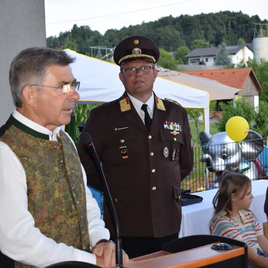 Ein Mann in weißem Hemd und Weste spricht an einem Podium, mit einem uniformierten Militäroffizier hinter ihm. Dahinter sitzt ein Mädchen an einem Tisch mit einer großen Elefantenstatue im Hintergrund.