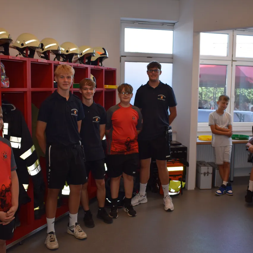 Sechs Jungen stehen in einer Feuerwache, tragen Uniformen und Helme. Ein Junge hält ein gelbes Objekt.