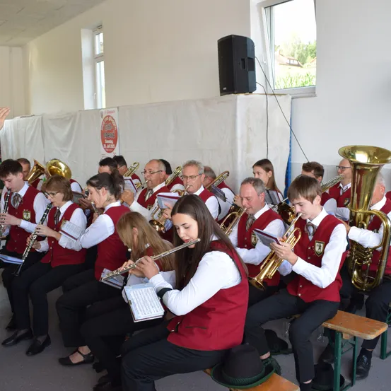 Eine Gruppe von Musikern in roten Westen sitzt und spielt Instrumente in einem Raum mit einem Lautsprecher an der Wand.