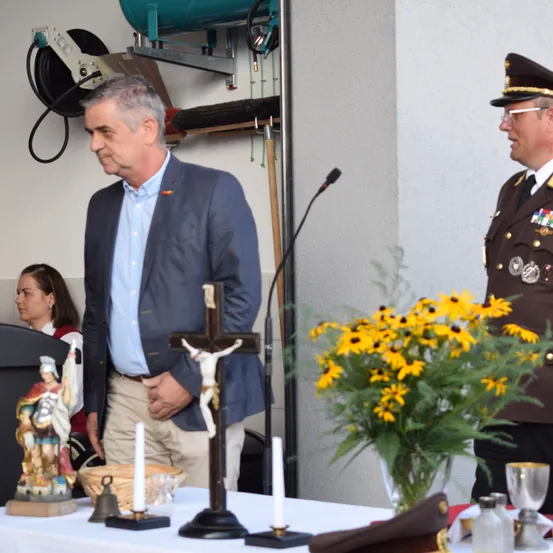 Ein Mann in einem blauen Anzug steht neben einem Tisch mit einem Kreuz, Kerzen und einem Strauß gelber Blumen. Ein uniformierter Offizier steht neben ihm. Eine Frau in einer roten Weste sitzt in der Nähe. Ausrüstung ist an der Wand hinter ihnen sichtbar.