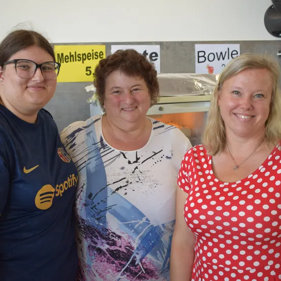 Drei Frauen lächeln und posieren für ein Foto. Die Frau auf der linken Seite trägt ein blaues Shirt mit einem Spotify-Logo. Die Frau in der Mitte trägt ein weißes Shirt mit einem abstrakten Design. Die Frau auf der rechten Seite trägt ein rotes Kleid mit Punkten. Hinter ihnen befindet sich ein gelbes Schild mit dem Wort 'Mehlspeise'.