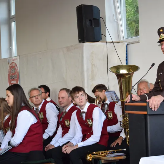 Eine Gruppe von Menschen in roten Westen sitzt in einer Reihe, möglicherweise in einer Kirche. Ein Mann in Uniform spricht in ein Mikrofon. Ein Lautsprecher und eine Tuba stehen davor.