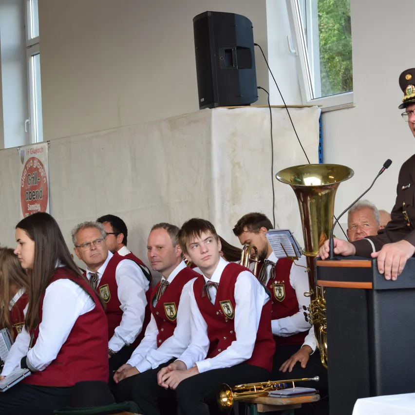 Eine Gruppe von Menschen in roten Westen sitzt in einer Reihe, möglicherweise in einer Kirche. Ein Mann in Uniform spricht in ein Mikrofon. Ein Lautsprecher und eine Tuba stehen davor.