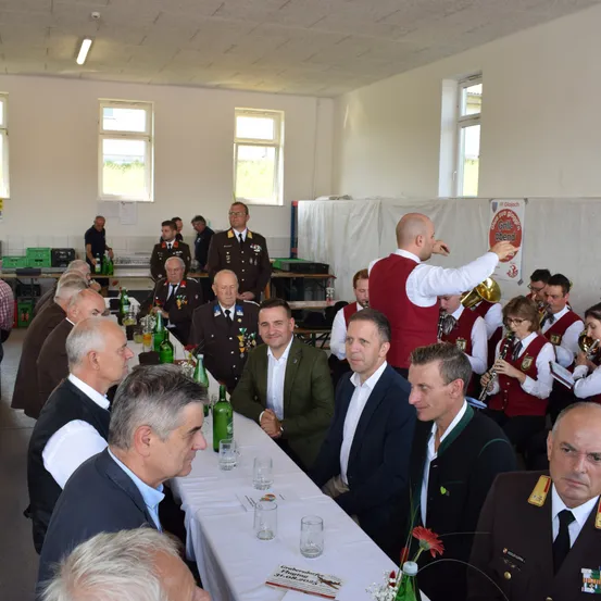 Mehrere Männer in formeller Kleidung sitzen um einen langen Tisch herum, während eine Band im Hintergrund Instrumente spielt. Ein Mann in einem roten Weste steht und deutet, während er eine Trompete hält. Der Raum ist gut beleuchtet mit natürlichem Licht aus den Fenstern.