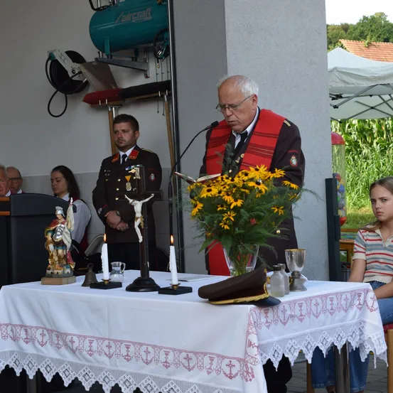 Ein Mann spricht an einem mit Blumen, einem Kruzifix und einem Hut geschmückten Podium, mit zwei brennenden Kerzen und einer Statue. Zwei Männer in Uniformen stehen hinter ihm, und andere sitzen im Hintergrund.