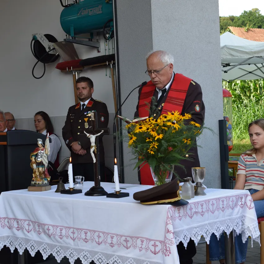 Ein Mann spricht an einem mit Blumen, einem Kruzifix und einem Hut geschmückten Podium, mit zwei brennenden Kerzen und einer Statue. Zwei Männer in Uniformen stehen hinter ihm, und andere sitzen im Hintergrund.