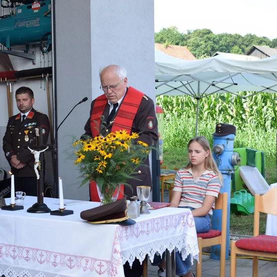 Ein älterer Mann spricht an einem Rednerpult mit einem Kreuz und Kerzen auf einem Tisch, ein Mädchen sitzt in der Nähe. Im Hintergrund ein Mann in Uniform und ein Maisfeld.