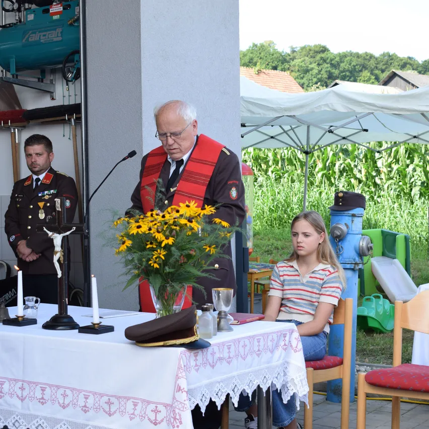 Ein älterer Mann spricht an einem Rednerpult mit einem Kreuz und Kerzen auf einem Tisch, ein Mädchen sitzt in der Nähe. Im Hintergrund ein Mann in Uniform und ein Maisfeld.