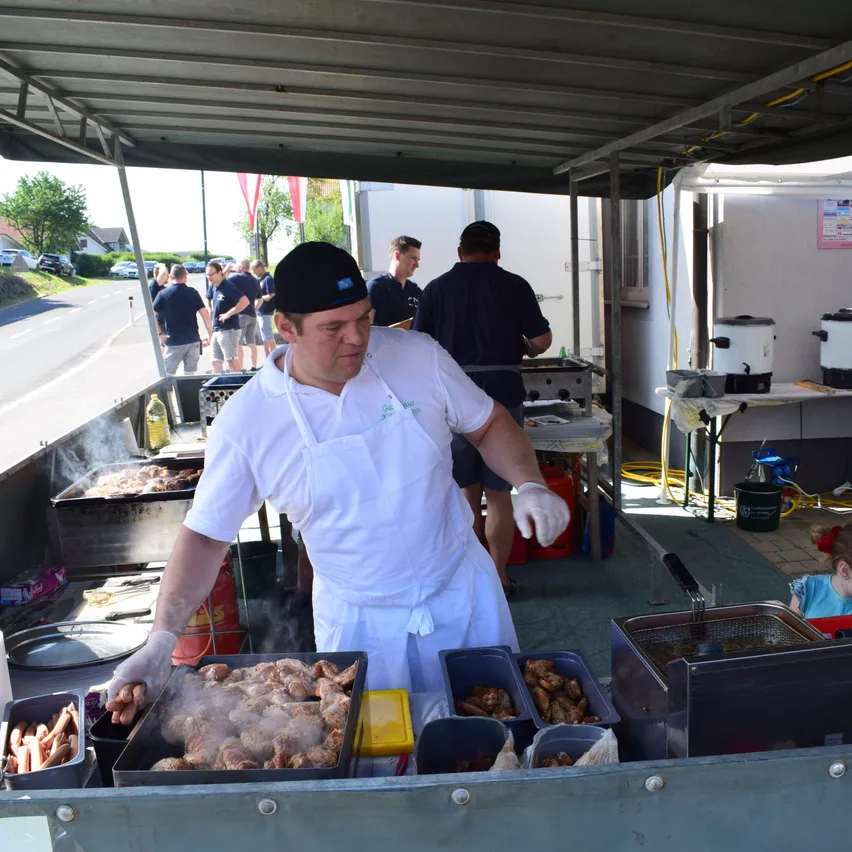 Ein Mann kocht Essen auf einem Grill unter einem Vordach. Mehrere Leute stehen hinter ihm. Es gibt verschiedene Kochutensilien und Behälter auf dem Grill. Bäume und Autos sind im Hintergrund zu sehen.