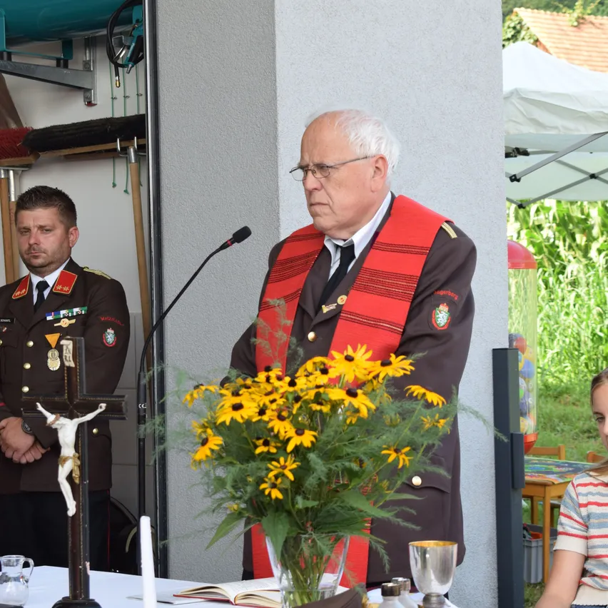 Ein älterer Mann steht hinter einem Podium mit einem Mikrofon, trägt eine rote und schwarze Schärpe und eine Brille. Zu seiner Linken steht ein Mann in Uniform neben einem Kruzifix. Ein Blumenarrangement steht auf dem Tisch vor ihnen.