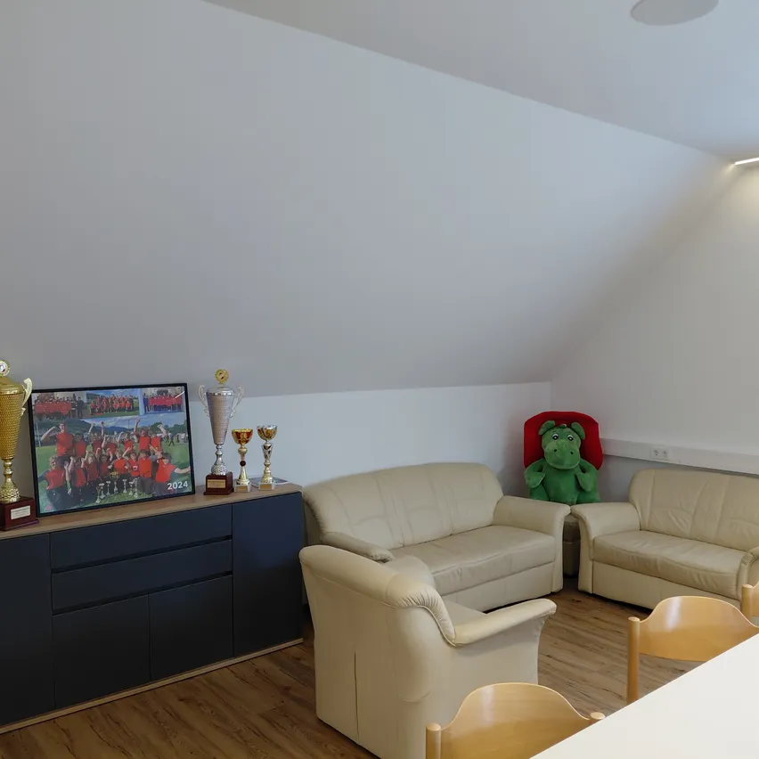 Ein Zimmer mit beigefarbenen Ledersofas, Holzstühlen, einem Schreibtisch, Trophäen und einem gerahmten Foto. Ein grünes Kuscheltier sitzt auf einem Sofa.