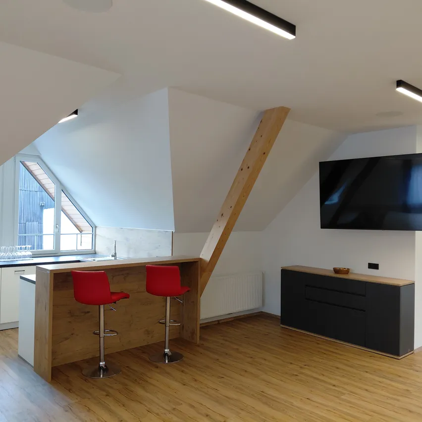 Ein Dachgeschosszimmer mit Holzboden und Küchenbereich. Zwei rote Hocker stehen vor einer Holztheke mit Waschbecken und Wasserhahn. Ein Fernseher ist an der Wand montiert. Ein Holzpfeiler steht in der Mitte des Raumes.
