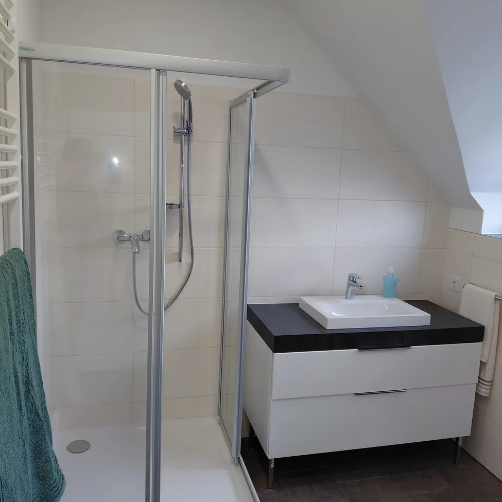Ein modernes Badezimmer mit einer Glasdusche, weißen Fliesenwänden und einem Waschtisch mit Waschbecken und Schubladen. Ein Handtuch hängt an einem Haken.