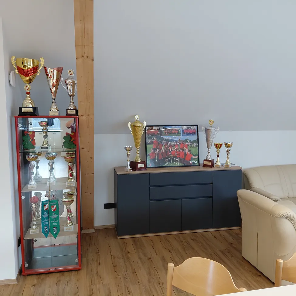 Ein Raum mit ausgestellten Trophäen, einem Foto auf einem Schrank, einer beigen Couch und zwei Stühlen. Der Boden ist aus Holz.