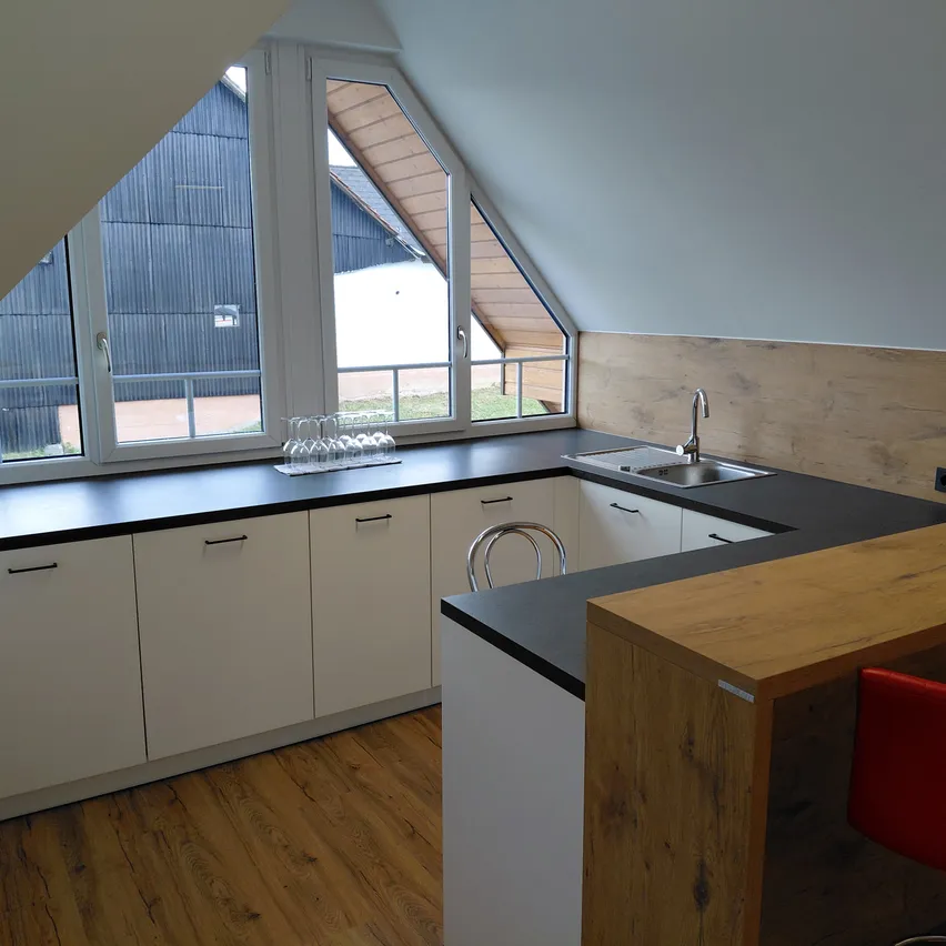 Eine Dachgeschossküche mit Holzböden, weißen Schränken, schwarzen Arbeitsplatten und einem Waschbecken mit Wasserhahn. Ein roter Stuhl steht rechts, und ein Fenster mit Blick auf ein Gebäude befindet sich links.