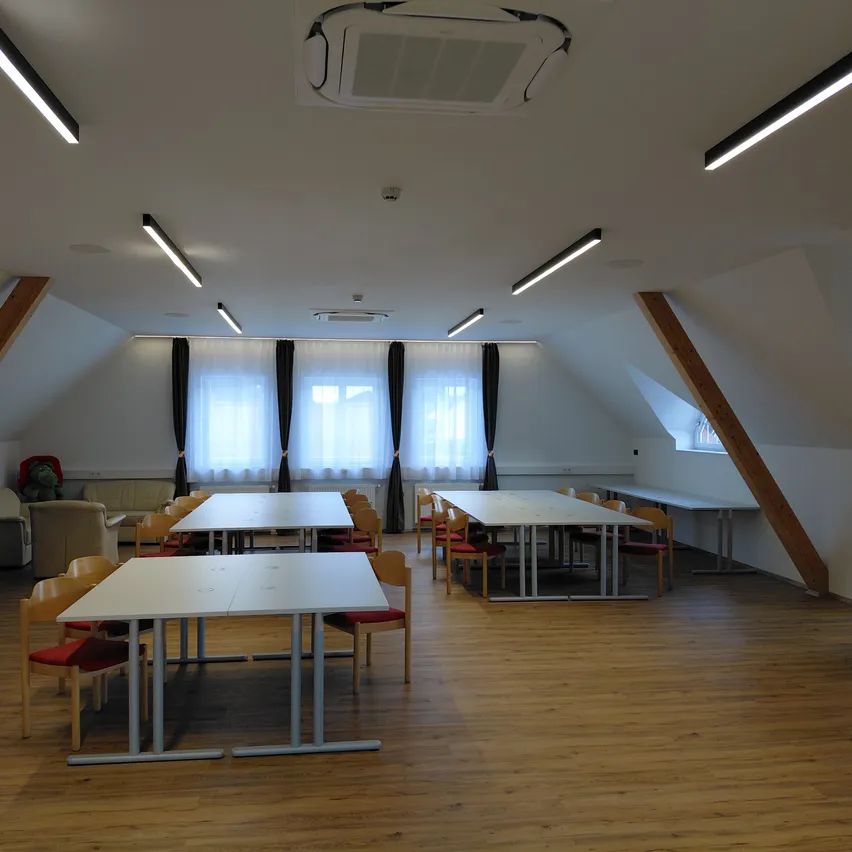 Ein leerer Sitzungssaal mit Holzboden, weißen Wänden und mehreren Tischen und Stühlen. Der Raum hat weiße Deckenleuchten und Fenster mit weißen Vorhängen.