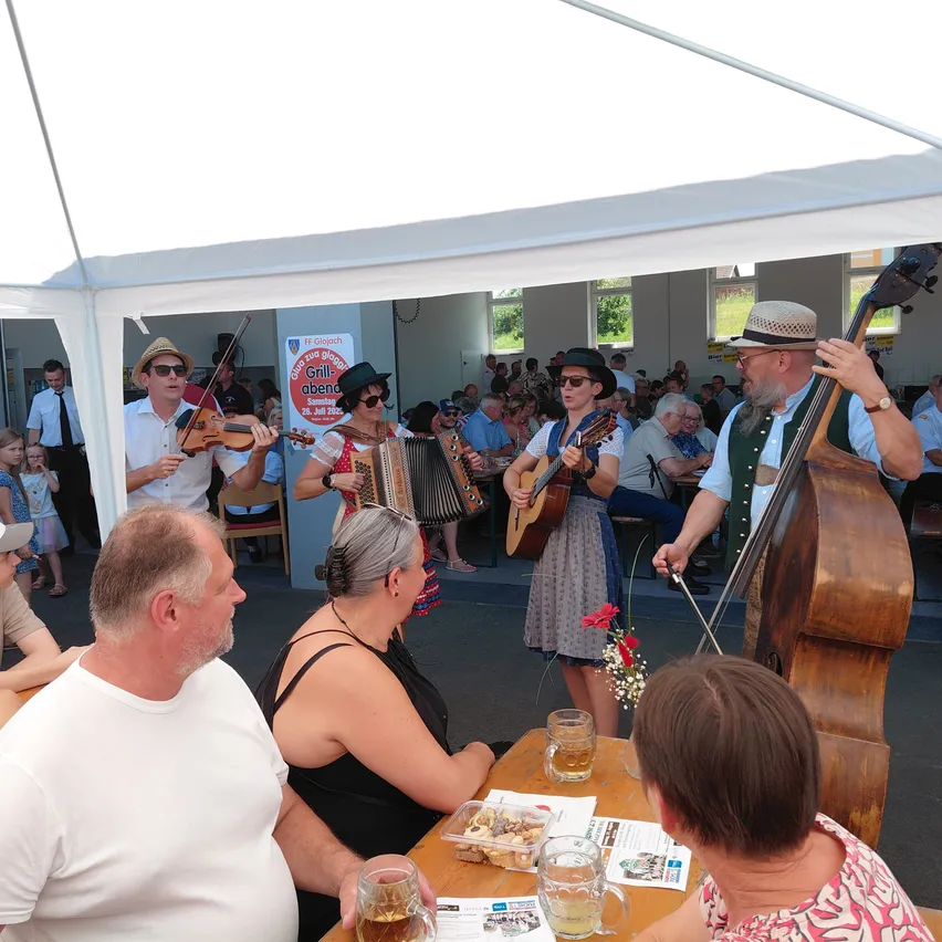 Eine Gruppe von Musikern tritt für ein Publikum unter einem weißen Zelt auf. Ein Mann spielt Cello, eine Frau Gitarre und eine andere Frau Akkordeon. Das Publikum sitzt und schaut zu.