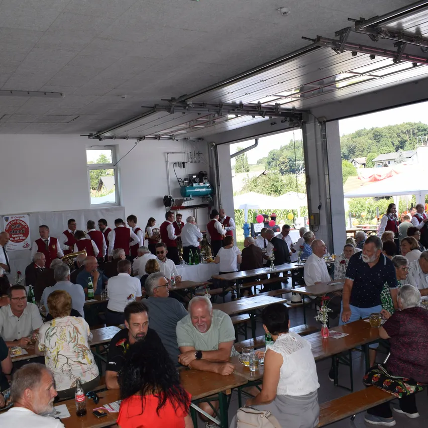 Eine große Gruppe von Menschen in roten Westen und weißen Hemden versammelt sich um Tische in einem Innenbereich. Die Umgebung umfasst lange Holztische, Stühle und einige stehende Personen. Der Raum hat große Fenster und eine Decke mit sichtbaren Lichtern. Draußen stehen einige Leute in der Nähe eines Zeltes.