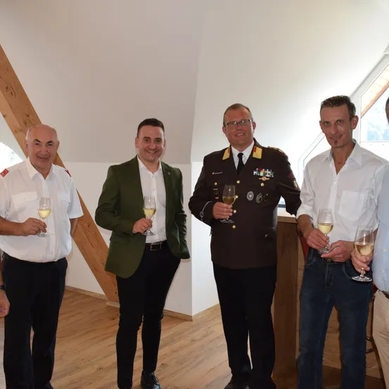 Fünf Männer stehen in einem Raum mit Holzboden und Balken, lächelnd und mit Weinglas in der Hand. Einer trägt eine Militäruniform, der andere eine grüne Jacke.