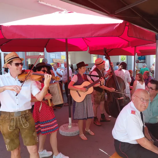 Eine Gruppe von Musikern in traditioneller bayerischer Kleidung spielt unter einem großen roten Regenschirm. Ein Mann spielt Geige, während eine Frau Akkordeon spielt. Eine andere Frau spielt Gitarre und ein Mann spielt Cello. Sie werden von einem älteren Mann begleitet, der auf einer Bank sitzt.