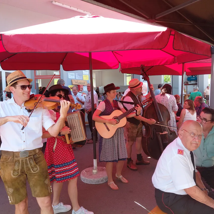 Eine Gruppe von Musikern in traditioneller bayerischer Kleidung spielt unter einem großen roten Regenschirm. Ein Mann spielt Geige, während eine Frau Akkordeon spielt. Eine andere Frau spielt Gitarre und ein Mann spielt Cello. Sie werden von einem älteren Mann begleitet, der auf einer Bank sitzt.