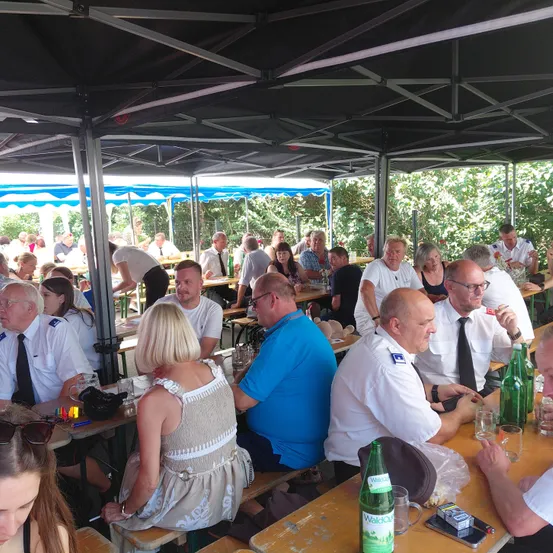 Eine Gruppe von Menschen sitzt unter einem Zelt an Tischen. Sie tragen Uniformen und einige haben Krawatten. Auf den Tischen stehen Flaschen und Gläser. Bäume sind im Hintergrund.